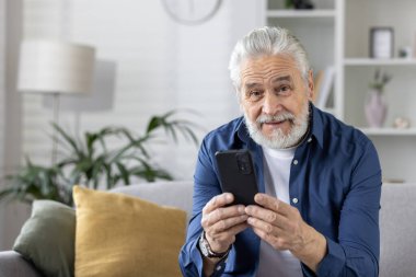 Gri sakallı neşeli yaşlı adam akıllı telefonuna bakarken gülümsüyor, evdeki kanepede rahatça oturuyor..