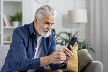 Neşeli, kıdemli bir adam rahat bir şekilde oturmuş, akıllı telefonuyla meşgul ve güzel ışıklandırılmış, rahat bir odada gülümsüyor..