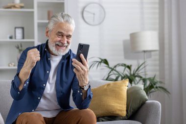 Beyaz saçlı neşeli olgun bir adam, akıllı telefonuna bakıp oturma odasında rahatça otururken sevinçle kutlama yapıyor..