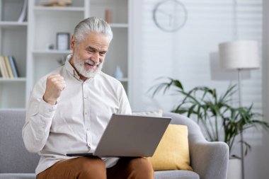 Yaşlı bir beyefendi dizüstü bilgisayar kullanıyor ve rahat bir oturma odasında zafer jesti yaparak heyecan gösteriyor..