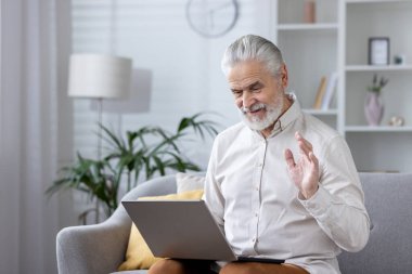 Yaşlı bir adam evde dizüstü bilgisayar kullanarak rahatlıyor ve modern bir oturma odasında dalgalı birini selamlıyor..