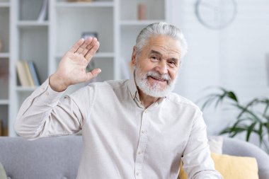 Neşeli yaşlı bir adam kameraya el sallıyor, sanki evdeki bir video görüşmesi gibi arkadaş canlısı ve meşgul görünüyor..