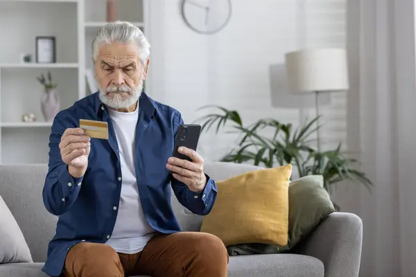 A mature adult male comfortably using a smartphone and credit card for a secure online transaction while sitting on a sofa at home.
