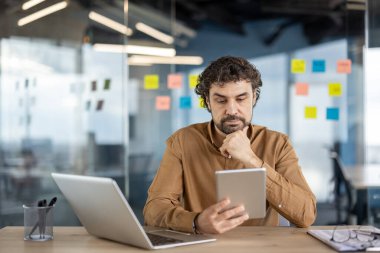 Modern ofis ortamında renkli yapışkan notlarla çevrili bir tableti elinde tutarken bir dizüstü bilgisayarda çalışan sıradan iş kıyafetleri içinde düşünceli bir adam..