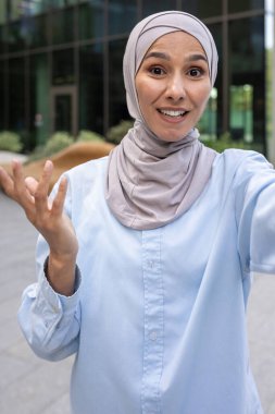 Mavi tesettürlü genç bir Müslüman kadının modern şehir arkaplanıyla selfie çektiği canlı bir fotoğraf..