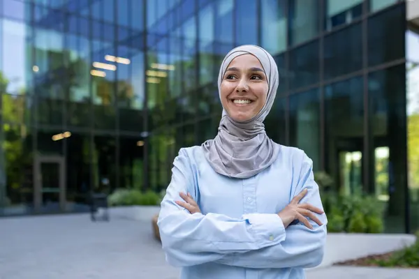 Professional Muslim woman in a hijab stands confidently outside an urban corporate building, portraying empowerment and diversity.