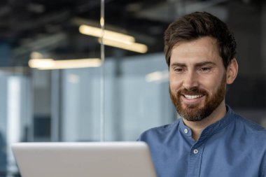 Ofiste mavi tişörtlü profesyonel bir erkek, dizüstü bilgisayarına bakarken gülümsüyor, özgüven ve pozitiflik yayıyor..