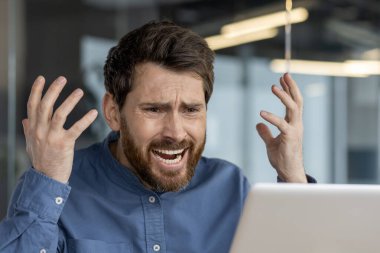 Modern ofis ortamında bilgisayar sorunlarıyla uğraşırken mavi gömlekli yetişkin bir erkek stresli ve sinirli hissediyor..