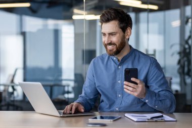 Bir dizüstü bilgisayar üzerinde çalışan ve modern ofis ortamında akıllı bir telefon tutan, üretkenliği ve bağlılığı ifade eden odaklanmış bir profesyonel..