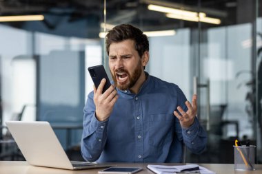 Modern ofis ortamında akıllı telefonuna bağıran genç bir erkek profesyonel, işle ilgili stres ve çatışmaları dile getiriyor..
