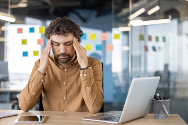 Kahverengi gömlekli bir erkek profesyonel, acı hisseder. Ofis masasında otururken başını tutar. Stres ya da migreni gösterir..