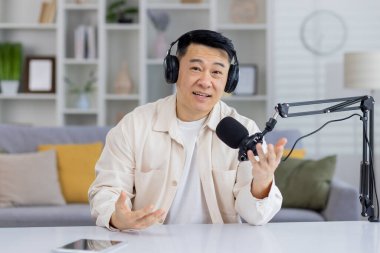 Dost canlısı, kulaklıklı Asyalı adam podcast yapıyor, kameraya doğru konuşuyor ve bir yayın mikrofonu ve ev stüdyosunda dizüstü bilgisayarla hareket ediyor..