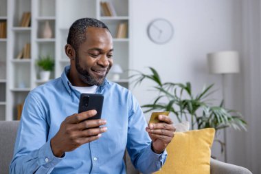 Çevrimiçi alışveriş için akıllı telefon ve kredi kartı kullanan sıradan bir adam evde rahat bir şekilde otururken iç mekanda ve parlak iç mekanlarda.