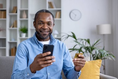 Akıllı bir telefon ve kredi kartı tutarken kameraya gülümseyen neşeli Afrikalı Amerikalı adam online alışveriş veya bankacılık portresi çiziyor..