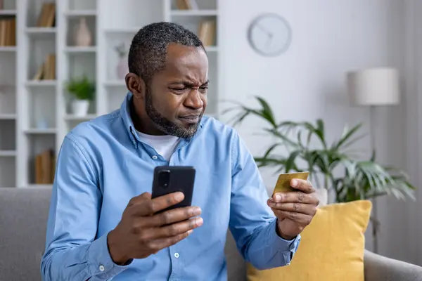 African American man experiences confusion and potential fraud while using his credit card and smartphone in a well-lit room.