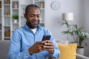 Mavi tişörtlü, akıllı telefonunu karıştırırken gülümseyen, aydınlık bir oturma odasında rahatça oturan olgun Afro-Amerikan bir adam..