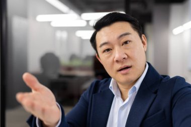 Ciddi Asyalı iş adamı video konferans sırasında cep telefonunun web kamerasına bakıyor ve elini kaldırıyor. Kendine güvenen erkek, sanal sohbetteki uzak çalışanlara acil soruları sormak için sesleniyor..