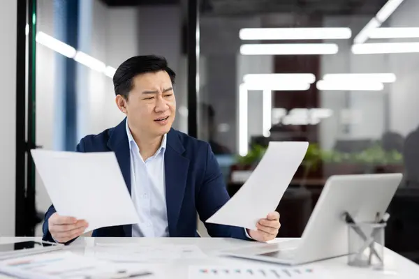 Puzzled chinese financial director looking amazingly in legal documents with counting in equipped cabinet. Nervous man in business suit trying figuring out solution to mistake of new employees.