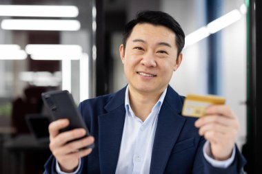 Takım elbiseli Asyalı bir işadamı akıllı telefon kullanır ve elinde bir kredi kartı tutarak mobil bankacılık, finans veya çevrimiçi alışveriş kavramını resmeder..
