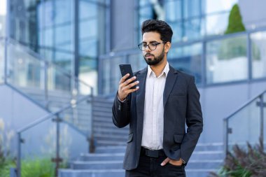 Takım elbiseli genç Hintli bir işadamı takım elbiseli bir ofis merkezinin yanında duruyor elini cebinde tutuyor ve telefonda bir video görüşmesi yapıyor..