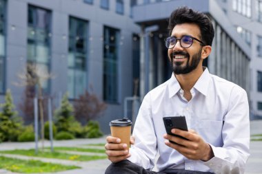 Beyaz gömlekli ve gözlüklü gülümseyen genç Hintli bir adam bir iş molasında, elinde telefon ve bir fincan kahveyle binanın dışında oturuyor ve mutlu bir şekilde yan tarafa bakıyor..