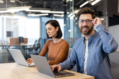 Bir erkek ve kadın yan yana çalışıyorlar, parlak, modern bir ofis ortamında, ikisi de dizüstü bilgisayar kullanıyor ve pozitif, odaklanmış bir tavır sergiliyorlar..