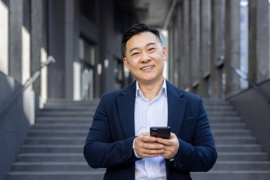 Ofis binasının dışında takım elbiseli genç bir Asyalı 'nın yakın plan portresi elinde telefonla kameraya gülümsüyor..