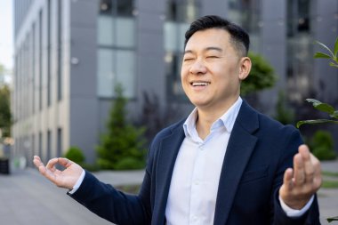 Close-up photo of young Asian male businessman sitting relaxed outside working office in lotus yoga pose and meditating with closed eyes. clipart