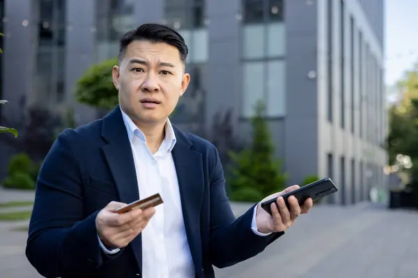Close-up portrait of young worried Asian man businessman sitting near office building, holding phone and credit card in hands and looking upset at camera, financial problems with account.