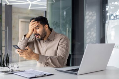 Hintli bir işadamı modern ofis ortamında akıllı telefonunu incelerken kafası karışık ve stresli görünüyor..