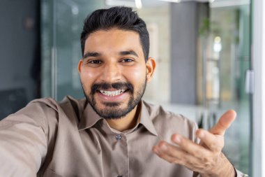 Gündelik gömlekli kendine güvenen iş adamı kameraya bakarken gülümsüyor ve telefonla konuşuyor, modern bir ofiste duruyor..