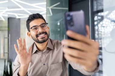 Çağdaş bir ofis ortamında akıllı telefonunu kullanan neşeli, profesyonel bir adam. Mutlulukla el sallıyor, pozitif enerji ve güven saçıyor..