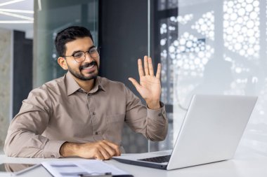 Akıllı gömlekli neşeli bir erkek profesyonel modern ofis ortamında sanal bir toplantı sırasında bilgisayarına sıcak bir şekilde el sallıyor..
