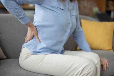 Son sınıf öğrencisi bir kadın kanepeye oturmuş, sırtını acıdan sımsıkı kavrıyor ve günlük hayattaki pek çok yaşlı tecrübenin verdiği rahatsızlığı vurguluyor..