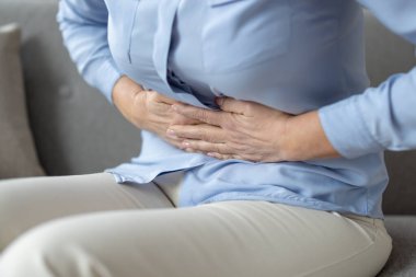 Son sınıf öğrencisi bir kadının kanepede otururken karnını tutarken görüntüsü. Resim rahatsızlığı ve yaşlanmanın zorluklarını betimliyor.