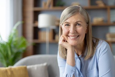 Neşeli, kıdemli bir kadının portresi ışıl ışıl gülümseyen, şık oturma odasında rahatça oturan, kendine güveni ve mutluluğu yayan.