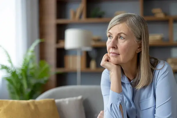 stock image A serene mature woman in a blue shirt stares thoughtfully while sitting in a well-lit living room. The peaceful atmosphere highlights her pensive mood.