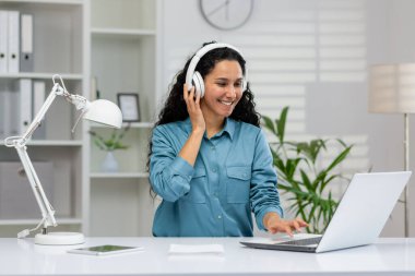 Mutlu kadın, dizüstü bilgisayarıyla iyi aydınlatılmış bir ofis ortamında çalışırken müzik dinlemeyi ve üretkenlik ve konfor göstermeyi seviyor..