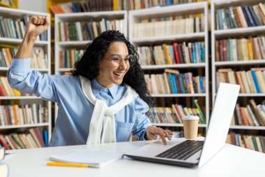 Kütüphanedeki neşeli bir kadın dizüstü bilgisayarı üzerinde çalışırken yumruğunu muzaffer bir şekilde kaldırıyor. Kitapların arasında neşe ve başarı yayar..