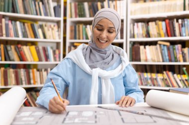 Neşeli bir Müslüman kadın mimar üniversite kütüphanesindeki bir masada bina planlarını çiziyor ve gözden geçiriyor. Başörtüsü takmış..