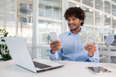 Modern ofis ortamında neşeli bir erkek girişimci elinde bir cep telefonu ve birden fazla Amerikan doları banknotuyla gülümsüyor ve telefona bakıyor..