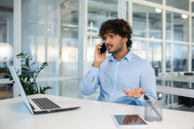 Genç İspanyol işadamı, modern bir ofis ortamında bilgisayarlı ve tabletli bir telefonla hayal kırıklığına uğramış ve kızgın görünüyor..