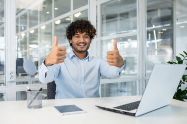 Açık mavi gömlekli genç bir adam parlak bir ofis ortamında bir masada oturuyor, hevesle iki parmağını kaldırıyor, gülümserken, işindeki başarı ve memnuniyeti resmediyor..