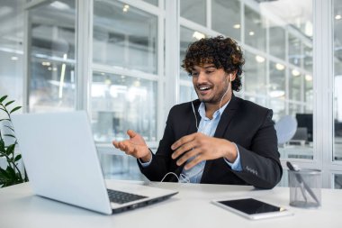 Dinamik profesyonel bir adam video görüşmesi yapmak için dizüstü bilgisayar ve kulaklık kullanır. Ofis ortamında cam duvarlar modern ve yoğun bir ortam sergiliyor..