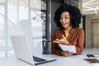 Kahverengi bluz giyen kıvırcık saçlı Afro-Amerikalı kadın, modern ofis ortamında elinde notlar tutarken aktif olarak bilgisayarını meşgul ediyor..