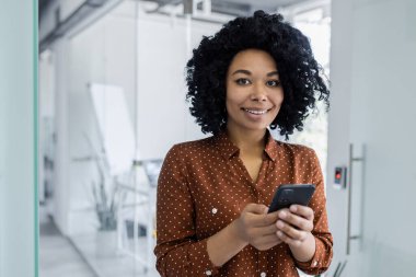 Kendine güvenen Afro-Amerikan iş kadını akıllı telefonunu güzel bir gülümsemeyle kullanıyor, parlak, modern bir ofiste duruyor. Profesyonel kıyafet ve pozitif işyeri enerjisi.