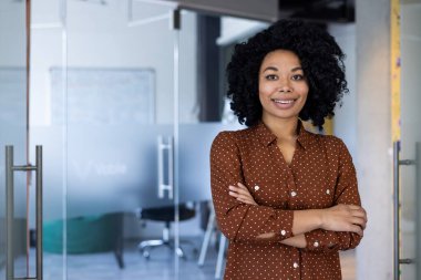 Kendine güvenen, kıvırcık saçlı, gülümseyen ve kollarını modern ofis ortamında kavuşturan genç bir kadının portresi. Profesyonelliği ve pozitifliği yansıtır..