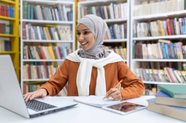 Kütüphanede tesettürlü neşeli bir Müslüman kadın, kitaplarla çevrili iken dizüstü bilgisayar kullanıyor. Notlar yazar, mutluluk saçar ve odaklanır..