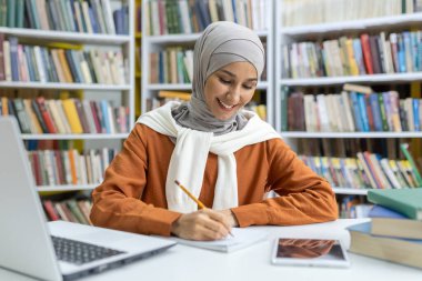 Kitaplarla çevrili, kütüphanede ders çalışan tesettürlü neşeli, genç bir Müslüman kadın. Not yazıyor ve bir dizüstü bilgisayar kullanıyor. Odaklanma ve mutluluk yayıyor..