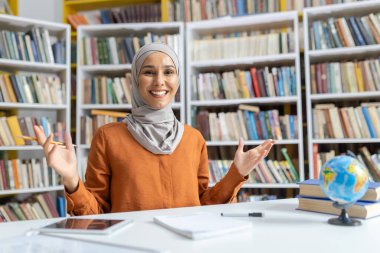 Kitaplarla dolu bir kütüphanede dururken kameraya gülümseyen tesettürlü, kendinden emin ve neşeli Müslüman kadın öğretmen..